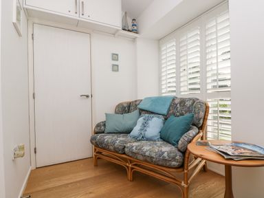 A small seating area with a floral patterned sofa with cushions next to a window with white blinds and a wooden side table with magazines at Sundowner in Padstow