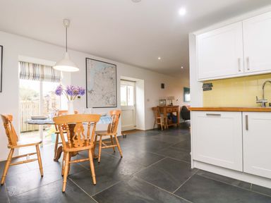 A dining area with wooden chairs and a table with flowers next to a kitchen with white cabinets and yellow tiles at Sundowner in Padstow