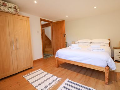 A bedroom with a wooden bed with white linens two striped rugs a wooden wardrobe and an open wooden door showing stairs at Lower Rissick Cottage in Sennen