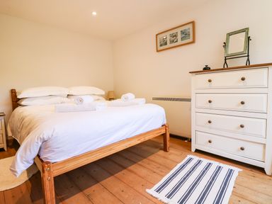 A bedroom with a wooden bed frame white bedding rolled towels on the bed a white chest of drawers with a mirror and framed pictures on the wall at Lower Rissick Cottage in Sennen