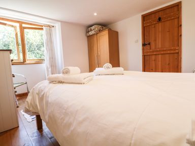 A bedroom with a bed with white sheets and towels a wooden wardrobe a wooden door and a window with curtains at Lower Rissick Cottage in Sennen