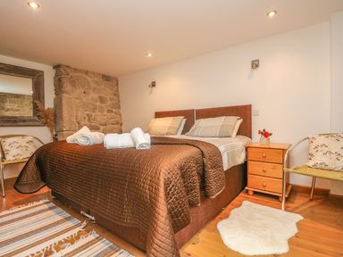 A bedroom with a double bed covered in a brown quilt and rolled white towels a wooden nightstand with a flower vase two chairs a striped rug a white fluffy rug and a stone wall at The Old Barn in Sennen