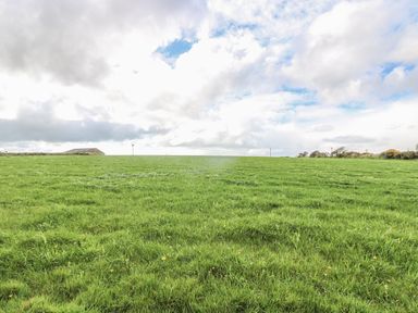 A green grassy field under a cloudy sky with a building and wind turbines in the distance at The Old Barn in Sennen