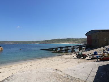 A beach with a boat ramp and a building with boats and a tractor at The Old Barn in Sennen