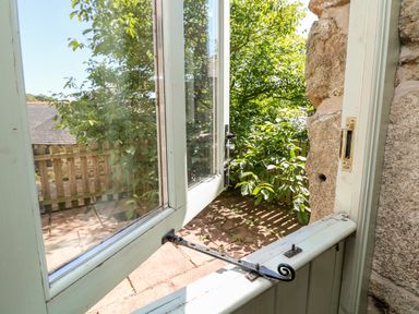 A partially open wooden window looking out onto a stone patio and green leafy trees outside at The Old Barn in Sennen