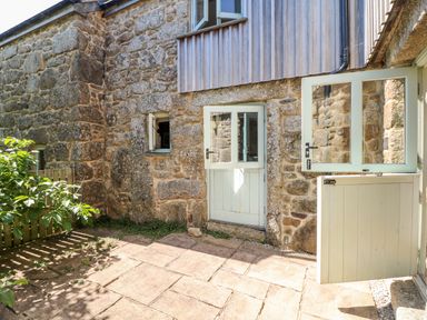 A stone exterior wall with windows and a half door opening to a paved courtyard at The Old Barn in Sennen
