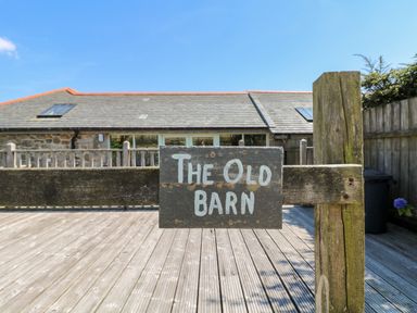 A wooden sign reading The Old Barn on a post with a stone building and wooden deck at The Old Barn in Sennen