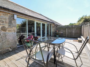 An outdoor wooden deck with a glass table and four metal chairs outside a stone building at The Old Barn in Sennen