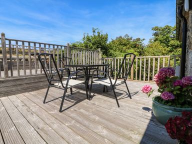 An outdoor wooden deck with a black metal table and four chairs and a planter with pink flowers at The Old Barn in Sennen
