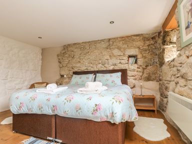 A bedroom with a bed with floral bedding a stone wall a wooden bedside table and a small window at Barneys Barn in Sennen