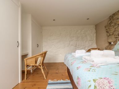 A bedroom with a floral bedspread wooden floor two wicker chairs white wardrobes and a stone wall at Barneys Barn in Sennen