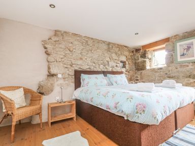 A bedroom with a double bed, wicker chair, small wooden side table, and stone walls at Barneys Barn in Sennen