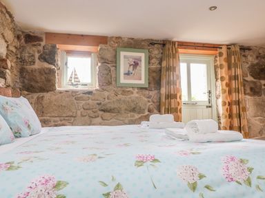 A bedroom with stone walls a bed with floral bedding folded towels on the bed a small window with a model sailboat a door with curtains at Barneys Barn in Sennen