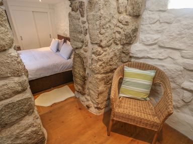 A bedroom with a bed and pillows visible through a stone archway and a wicker chair with a striped cushion in the corner at Barneys Barn in Sennen