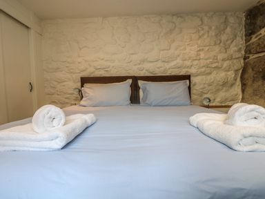 A bed with light blue sheets and two pillows in front of a white stone wall with rolled white towels on the bed at Barneys Barn in Sennen