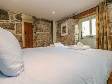 A bedroom with a bed made with white linens stone walls a wooden door and a window with curtains at Barneys Barn in Sennen
