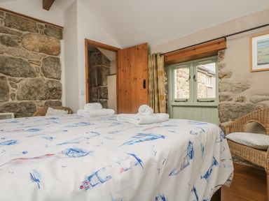 A bedroom with a bed with lighthouse patterned bedding a wicker chair stone walls a wooden door and a window at Barneys Barn in Sennen