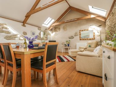 A dining area with wooden table and chairs next to a living area with beige sofas and wooden beams on the ceiling at Barneys Barn in Sennen