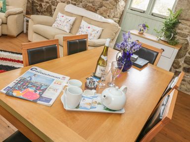 A dining table with a teapot cups wine bottle glasses newspapers and a flower vase in a living room with sofas and a sideboard at Barneys Barn in Sennen