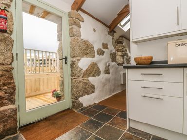 A kitchen area with white cabinets and stone and plaster walls next to a glass door leading outside at Barneys Barn in Sennen