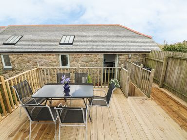 An outdoor wooden deck with a glass table and six chairs next to a stone building at Barneys Barn in Sennen