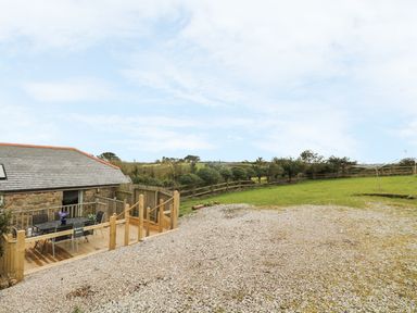 An outdoor area with a wooden deck and table with chairs next to a stone building and a gravel surface with grass and trees at Barneys Barn in Sennen