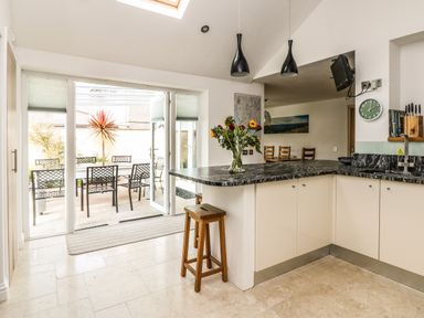 A kitchen with black countertop and wooden stools overlooking an outdoor dining area with metal chairs at Ridgeback House in Mullion
