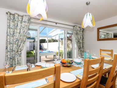 A dining area with a wooden table set for six people near sliding glass doors with patterned curtains showing a pool table outside at Ridgeback House in Mullion