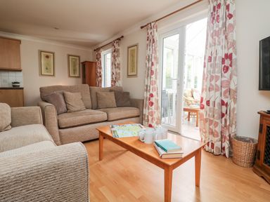 A living room with beige sofas a wooden coffee table with books and mugs curtains with floral patterns and glass doors at Tre Pol Pen in Falmouth