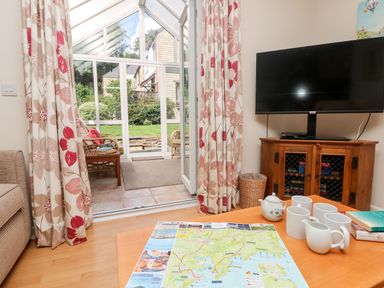 A living room with a coffee table holding a map and tea set a television on a wooden cabinet and a glass door leading to a sunroom with wicker chairs and floral curtains at Tre Pol Pen in Falmouth