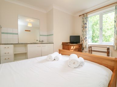 A bedroom with a wooden bed, rolled white towels on the bed, a window with floral curtains, a wooden chest of drawers, a small TV, and a sink with a mirror at Tre Pol Pen in Falmouth