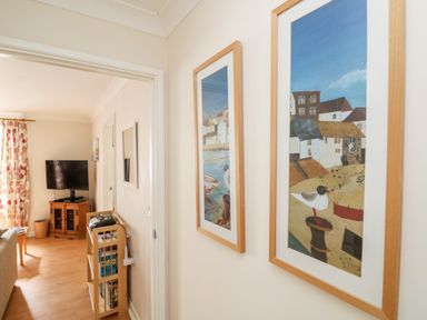 A hallway with framed artwork on the wall and a wooden bookshelf beside a living room with a television and floral curtains at Tre Pol Pen in Falmouth