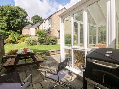 A patio with a wooden picnic table set with dishes two chairs and a barbecue next to a glass conservatory at Tre Pol Pen in Falmouth