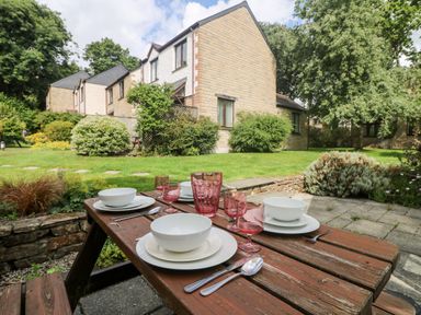 A wooden outdoor table set with white plates bowls cutlery and pink glasses in a garden area with grass bushes and stone houses at Tre Pol Pen in Falmouth