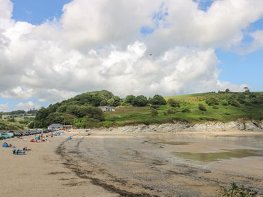 A sandy beach with people and parked cars near a green hillside under a partly cloudy sky at Tre Pol Pen in Falmouth