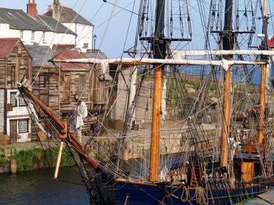 A docked wooden sailing ship with rigging near old wooden buildings and white houses by the water at Speedwell in Charlestown
