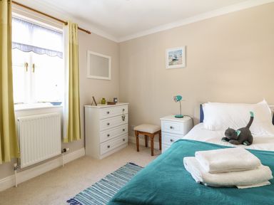 A bedroom with a bed with white sheets and a stuffed cat on it a teal blanket with folded towels a white nightstand with a lamp a white dresser a small stool a window with green curtains and a blue striped rug at Speedwell in Charlestown