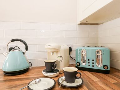 A kitchen countertop with a blue kettle, a white coffee machine, a blue toaster, two black cups with saucers, and two spoons at Speedwell in Charlestown