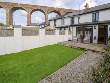 A backyard with green lawn stone patio with chairs white house and stone arch bridge in the background at Anneth Lowen in Angarrack near Hayle