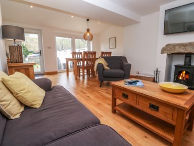 A living room with a sofa, armchair, wooden coffee table, dining table with chairs, lamp, wood-burning stove, and television at Anneth Lowen Angarrack near Hayle
