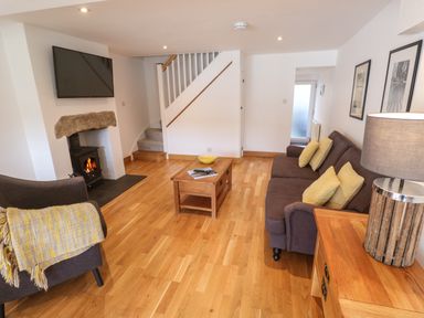 A living room with a wood burning stove a mounted TV a brown sofa with yellow cushions a wooden coffee table and a wooden sideboard with a lamp at Anneth Lowen in Angarrack near Hayle