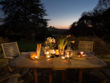 An outdoor wooden table with chairs set with wine bottles glasses lanterns flowers bread and cheese at Nairn in Braunton