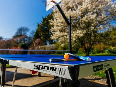 A blue table tennis table with paddles and an orange ball outdoors near a basketball hoop at Nairn in Braunton