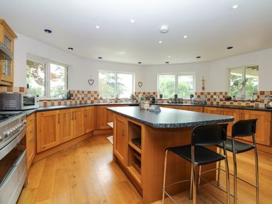 A kitchen with wooden cabinets a central island with two black chairs and windows overlooking trees at Nairn in Braunton