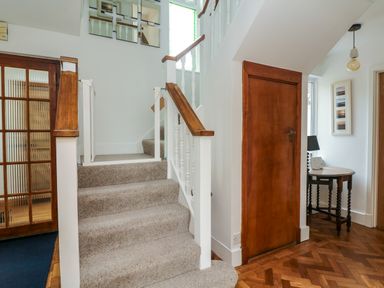 A staircase with carpet and wooden banisters beside a wooden door and a hallway with a table and lamp at Nairn in Braunton