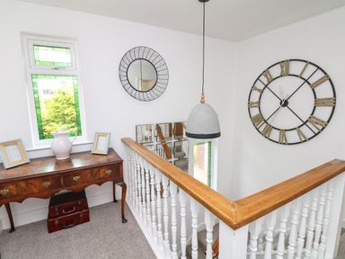 A stairway landing with a wooden desk holding a pitcher and two picture frames a round wall mirror a large clock and a hanging light at Nairn in Braunton