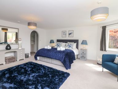 A bedroom with a large bed with blue bedding and pillows a blue rug a blue armchair a dresser with a mirror and two bedside tables with lamps at Nairn in Braunton