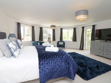 A bedroom with a large bed with pillows and a blue blanket two blue chairs a gray dresser with a television and windows with dark curtains at Nairn in Braunton