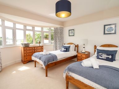 A bedroom with two single wooden beds blue throws and white towels a wooden chest of drawers and nightstand under a bay window at Nairn in Braunton