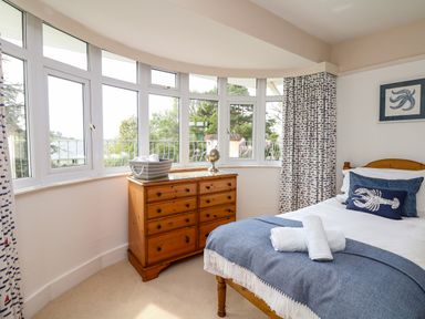 A bedroom with a wooden chest of drawers a bed with blue and white bedding and large windows with patterned curtains at Nairn in Braunton